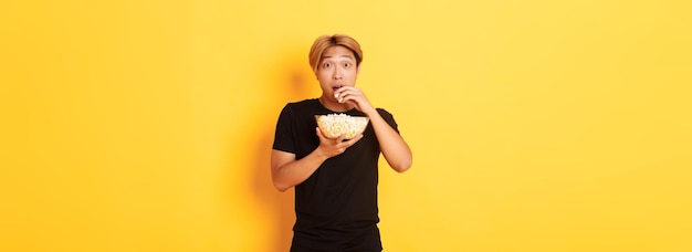 Un asiatique excité et étonné regarde un film intéressant et mange du pop-corn dans un bol jaune debout