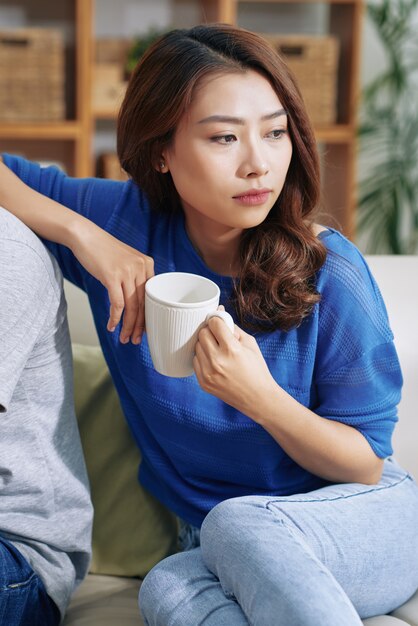 Asiatique femme assise sur un canapé à la maison avec une tasse et s'appuyant sur le dos d'un homme méconnaissable