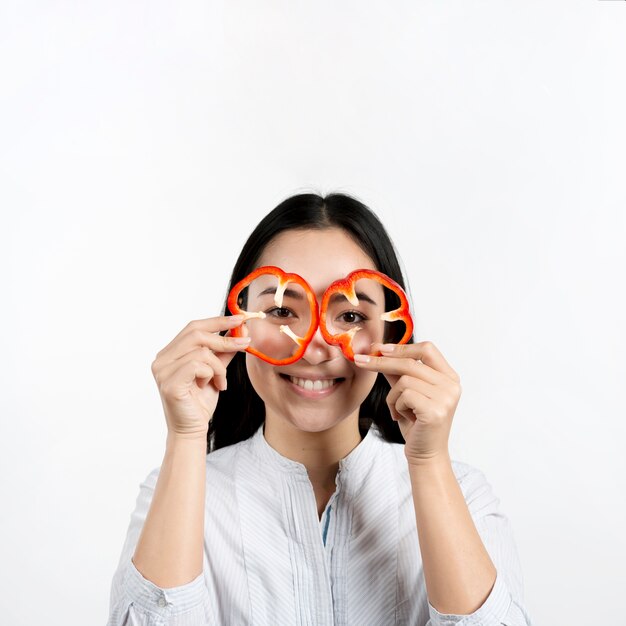 Asiatique femme tenant deux tranches de poivrons rouges devant ses yeux au-dessus de la surface blanche