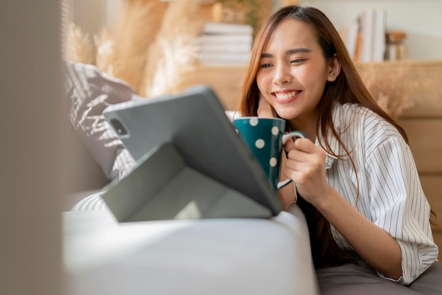 Asiatique séduisante femme porter un tissu décontracté maison isoler la quarantaine profiter de regarder le programme de films en ligne bradcast en streaming via une tablette sur le sol à côté du canapé dans le salon intérieur de la maison fond confortable