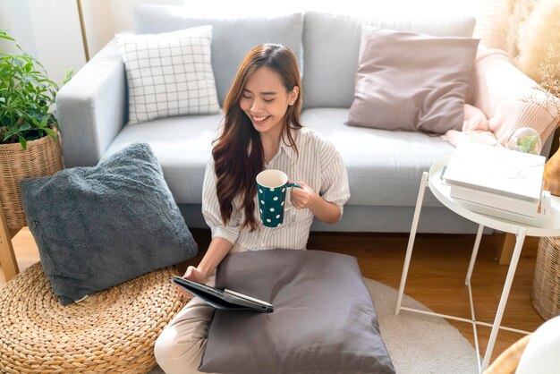 Asiatique séduisante femme porter un tissu décontracté maison isoler la quarantaine profiter de regarder le programme de films en ligne bradcast en streaming via une tablette sur le sol à côté du canapé dans le salon intérieur de la maison fond confortable