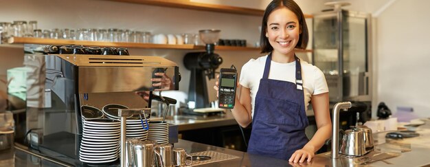 Une asiatique souriante, bariste, propriétaire d'un café en tablier, montrant un lecteur de cartes de paiement.