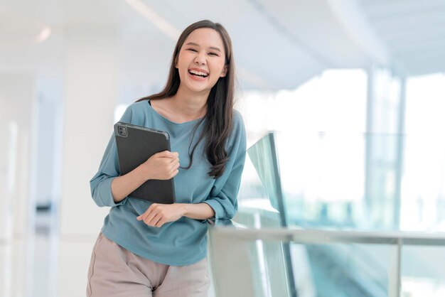 Asiatique souriante gaie femme nomade numérique main tenir la tablette regarder la caméra portrait shotbonheur souriant asie femme debout dans le couloir de l'université de bureau avec une attitude souriante positive