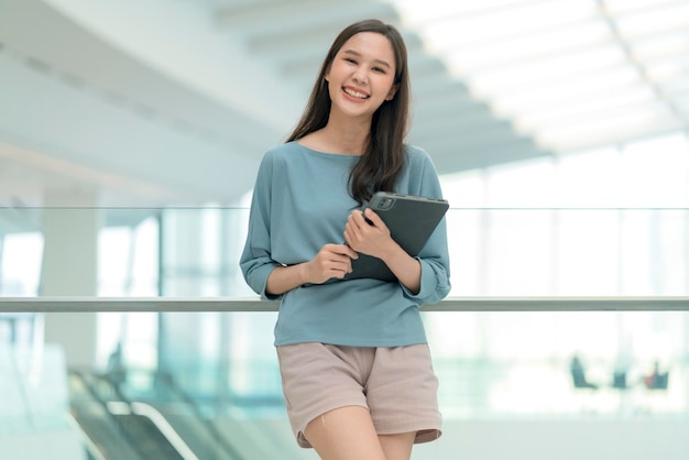 Asiatique souriante gaie femme nomade numérique main tenir la tablette regarder la caméra portrait shotbonheur souriant asie femme debout dans le couloir de l'université de bureau avec une attitude souriante positive