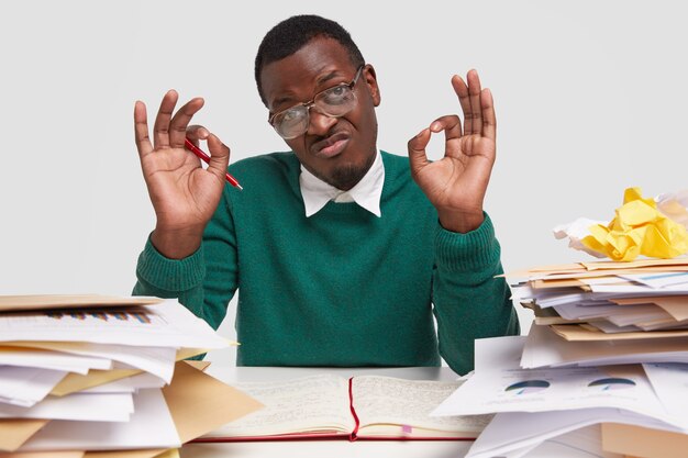 Assertif jeune homme à la peau foncée, porte des lunettes, un pull vert, fait un geste correct, approuve qu'il terminera le travail à temps