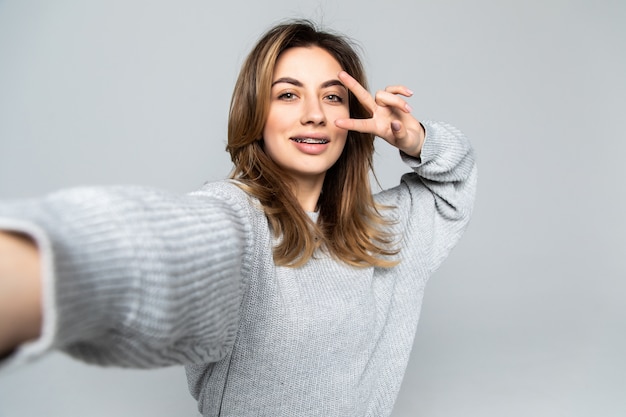 Assez gaie femme brune montrant un geste de paix tout en prenant selfie sur smartphone, isolé sur mur gris