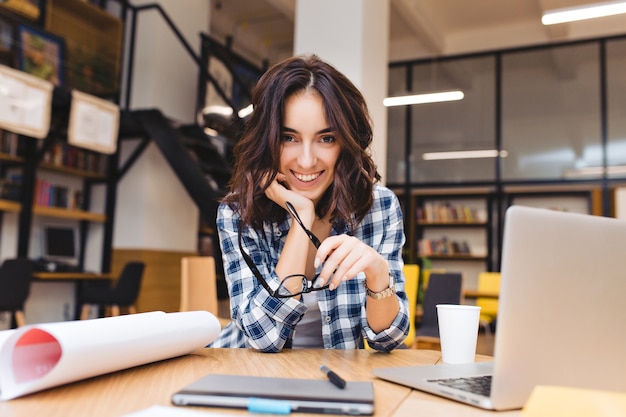 Assez joyeuse jeune femme assise sur la table des trucs de travail. Sourire, concevoir, travailler comme pigiste, vie étudiante, bonne humeur, carrière, grand succès.