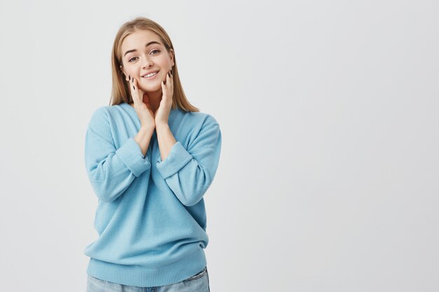 Assez souriante jeune belle femme blonde regardant avec des yeux calmes étant heureux d'entendre de bonnes nouvelles. Belle fille regardant toucher ses joues, posant en studio.