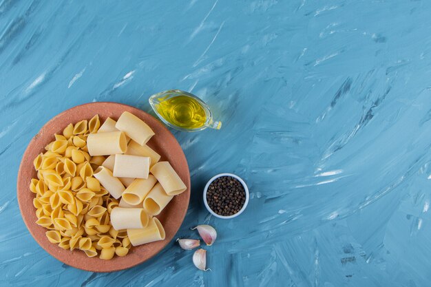 Une assiette en argile de pâtes non cuites avec de l'huile et des tomates rouges fraîches sur fond bleu.