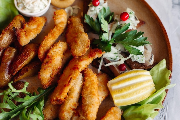 Assiette d'argile pleine d'apéritifs servis avec des pépites de poulet dorées cuites avec une croûte croustillante de délicieux canapés avec du hareng et des tomates cerises sauce à l'ail décorées de salade fraîche et de fromage