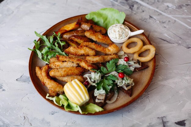 Assiette en argile pleine d'apéritifs, y compris des pépites de poulet dorées avec une croûte croustillante, de délicieux canapés avec du hareng et des tomates cerises servis avec une sauce à l'ail décorée de feuilles de salade et de fromage