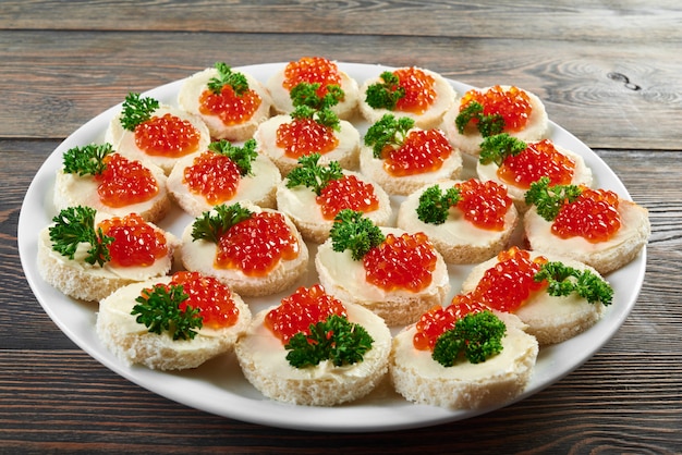 Assiette blanche sur la table en bois, pleine de petits canapés au beurre, caviar rouge et décorée de feuilles de persil vert frais. Apéritif savoureux contre restauration alcoolique ou buffet de restaurant