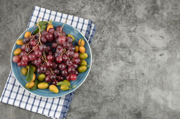 Assiette bleue de fruits kumquat et raisins rouges sur une surface en marbre.