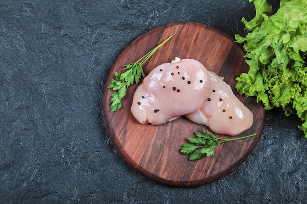 Assiette en bois de filet de poulet cru avec des verts sur table sombre. Vue de dessus.