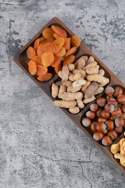 Assiette en bois pleine de diverses noix, craquelins et abricots secs sur une surface en marbre.