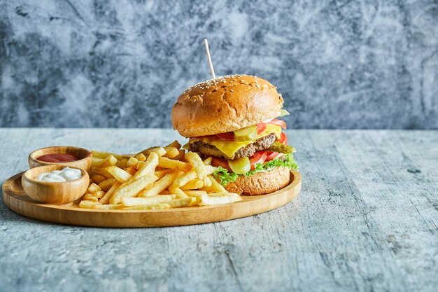 Une assiette en bois pleine de hamburger, pommes de terre frites avec du ketchup et de la mayonnaise sur la table en marbre.