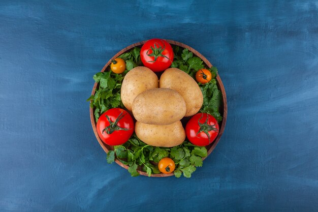 Assiette en bois de pommes de terre fraîches et tomates sur une surface bleue.