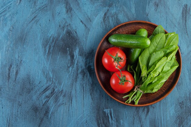 Assiette en bois de tomates fraîches, concombres et verts sur fond bleu.