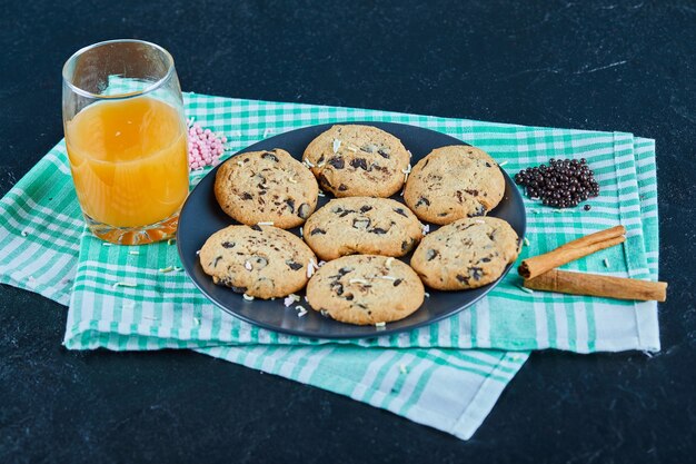 Une assiette de cookies aux pépites de chocolat et un verre de jus d'orange sur table noire avec de la cannelle