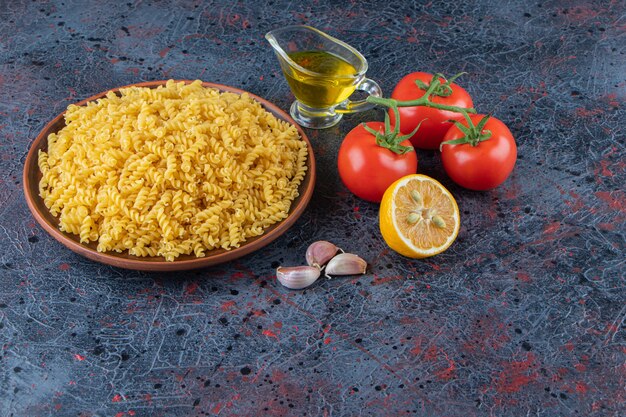 Une assiette de pâtes en spirale non cuites avec de l'huile et des tomates rouges fraîches sur un fond sombre.