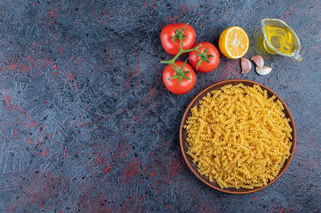 Une assiette de pâtes en spirale non cuites avec de l'huile et des tomates rouges fraîches sur un fond sombre.