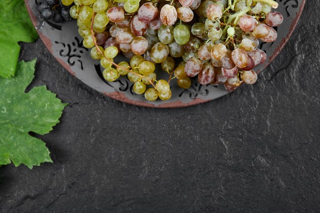 Une assiette de raisins mélangés avec des feuilles sur fond sombre. Photo de haute qualité