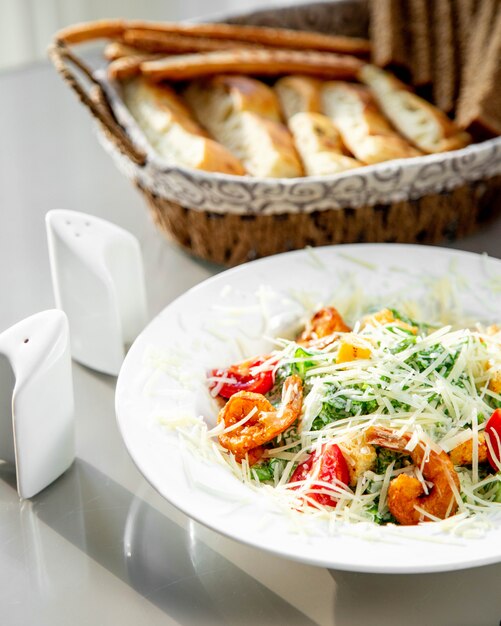 Une assiette de salade César aux crevettes servie avec une corbeille à pain, du sel et du poivre