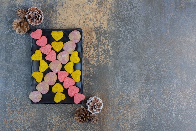 Photo gratuite une assiette sombre pleine de bonbons à la gelée sucrée en forme de cœur avec des pommes de pin. photo de haute qualité