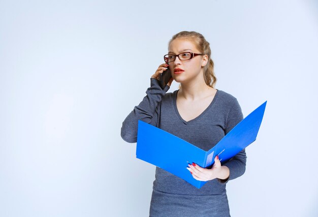 Assistant avec un dossier bleu parlant au téléphone et semble stressé car quelque chose ne va pas.