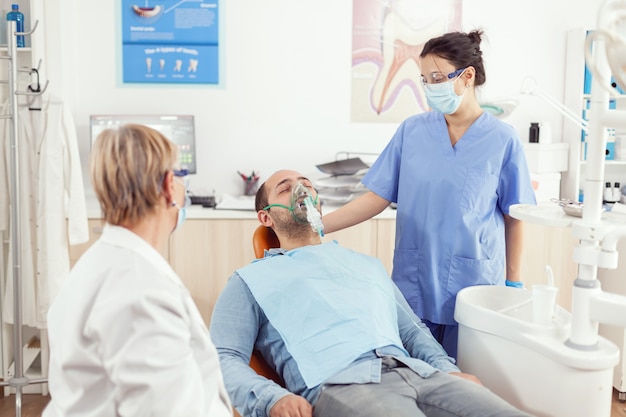 Assistant hospitalier mettant un masque à oxygène à un patient malade après une chirurgie de stomatologie, assis sur un fauteuil dentaire dans une chambre d'hôpital orthodontique pendant une consultation médicale Médecin dentiste examinant un mal de dents