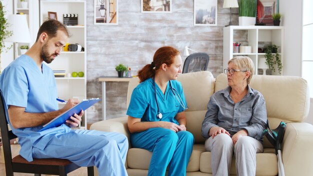 Assistant masculin prenant des notes sur le presse-papiers pendant qu'une femme médecin parle avec une femme âgée dans une maison de soins infirmiers