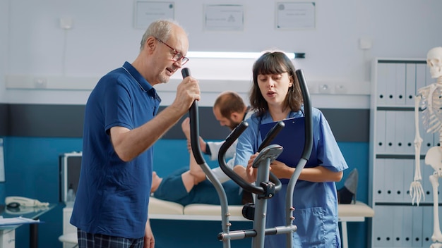 Assistant médical soutenant un homme âgé faisant de la physiothérapie, faisant de l'exercice sur un vélo stationnaire. Infirmière aidant une personne âgée à assister à la physiothérapie de réadaptation dans le cabinet.