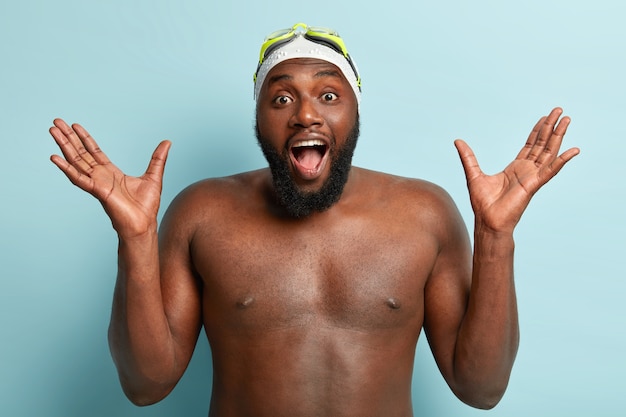 Photo gratuite athelitc nageur à la peau sombre avec des poils épais, lève les mains, pose nu contre un mur bleu, porte un bonnet de bain et des lunettes, a une formation de gymnastique, s'exclame des émotions positives.