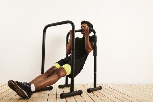 athlète afro-américain musclé en tenue d'entraînement noir faisant des rangées de poids corporel sur des barres mobiles contre un mur blanc et un plancher en bois clair.
