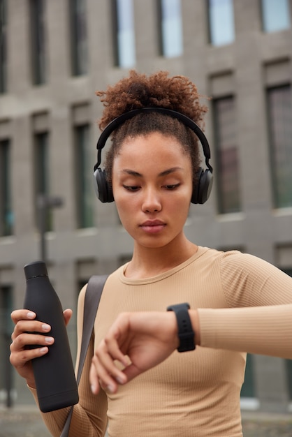 Une athlète afro-américaine boit de l'eau douce après l'entraînement vérifie le rythme cardiaque sur smartwatch écoute de la musique dans des poses d'écouteurs en ville