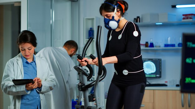 Athlète féminine avec masque courant sur cross trainer dans un laboratoire de sciences du sport mesurant les performances et la consommation d'oxygène pendant le test vo2max. chercheur médical regardant le balayage d'ecg surveillant la fréquence cardiaque