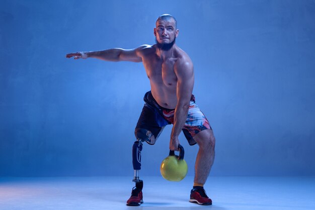 Athlète handicapé ou amputé isolé sur mur bleu. Sportif masculin professionnel avec formation de prothèse de jambe avec poids en néon. Sport handicapé et dépassement, concept de bien-être.