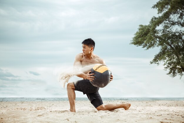 Athlète jeune homme en bonne santé, faire de l'exercice avec ballon à la plage. Signle modèle masculin air d'entraînement torse nu au bord de la rivière en journée ensoleillée. Concept de mode de vie sain, sport, fitness, musculation.