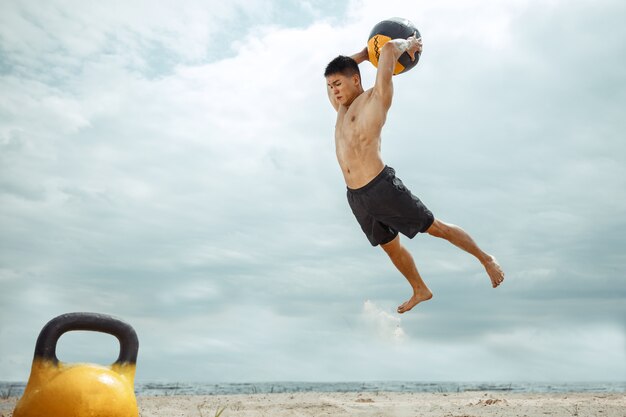 Photo gratuite athlète jeune homme en bonne santé, faire de l'exercice avec le poids et le ballon à la plage. signle modèle masculin formation torse nu au bord de la rivière. concept de mode de vie sain, sport, fitness, musculation.