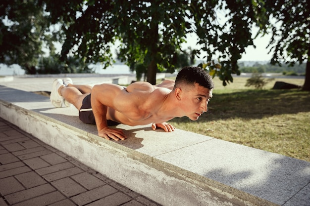 Un athlète masculin musclé faisant de l'exercice dans le parc. Gymnastique, entraînement, flexibilité des entraînements de fitness. Ville d'été en journée ensoleillée sur le terrain de fond. Mode de vie actif et sain, jeunesse, musculation.