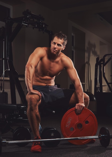 Un athlète musclé torse nu tient un disque d'haltères tout en étant assis sur un banc dans la salle de sport.