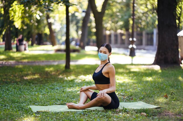 Athlétique jeune femme dans un masque de protection médicale, faire du yoga dans le parc le matin, formation des femmes sur un tapis de yoga