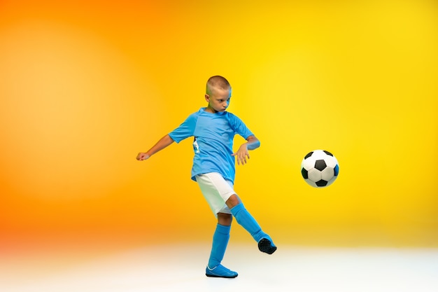 Attaque. Jeune garçon en tant que joueur de football ou de football en vêtements de sport pratiquant sur un dégradé jaune à la lumière du néon