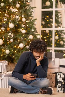 En attendant les vacances. jeune homme indien adulte en pull bleu et jeans regardant un smartphone touchant la main à face assis sur le sol près de l'arbre de noël et des cadeaux