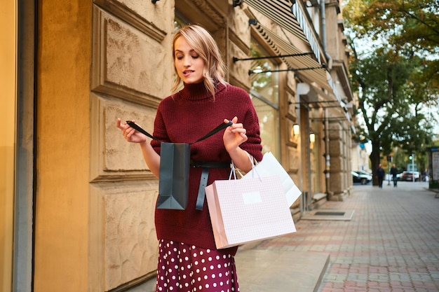 Attractice fille blonde styliah excitée regardant pensivement dans un sac à provisions sur la rue de la ville