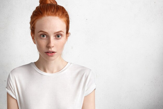 Attractive belle jeune femme au gingembre avec une expression sérieuse, a la peau de taches de rousseur, habillée avec désinvolture, pose contre l'espace de copie vierge