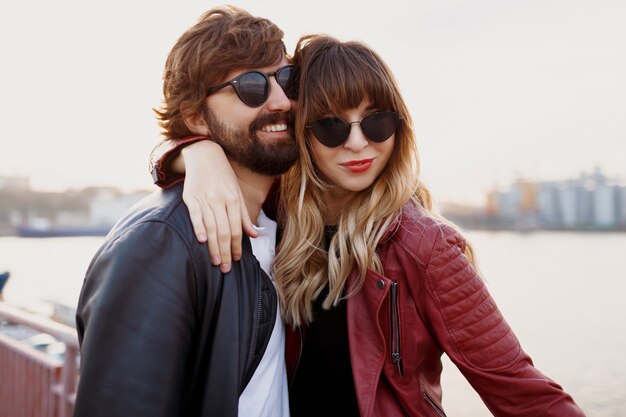 Attractive couple élégant amoureux posant en plein air, étreignant et marchant sur le quai. Couleurs douces du soir. Look à la mode. Lunettes de soleil tendance. Homme et femme embarrassants.