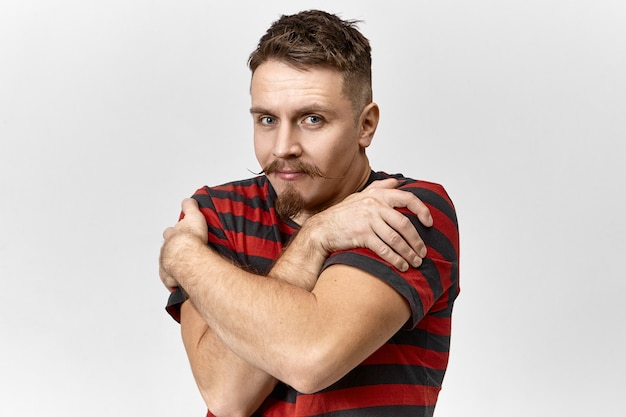 Attractive jeune homme barbu musclé portant un t-shirt rayé croisant les bras sur sa poitrine, gardant les mains sur les épaules comme si elle avait froid. Beau mec avec moustache s'embrassant en studio