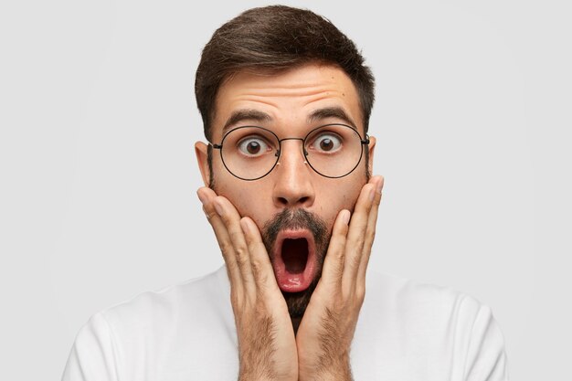 Attractive jeune homme avec une expression stupéfaite, touche les joues avec les deux paumes, regarde la caméra, ayant peur de la phobie