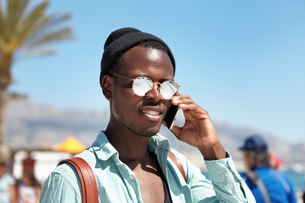 Attrayant étudiant afro-américain à la mode passant des appels téléphoniques à ses parents, partageant des nouvelles et des impressions tout en voyageant seul dans un pays étranger